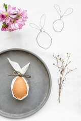 Ein braunes Ei mit weißen Hasenohren auf einem grauen Teller. Weißes Tischtuch, Draufsicht, basteln.