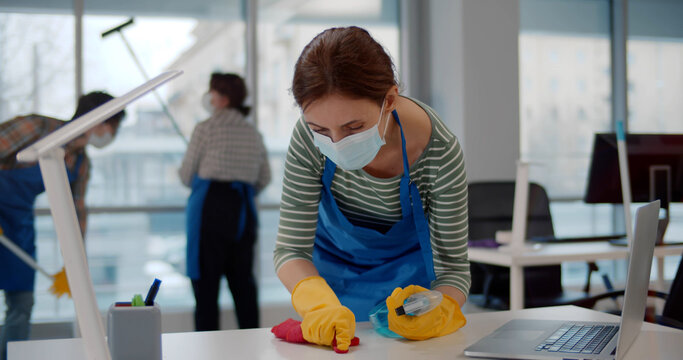 Portrait of young female worker in uniform and safety mask cleaning desk with rag and spray in office