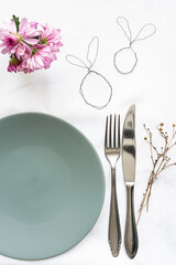 Ein leerer mintfarbener Teller mit Besteck und kleinen Osterhasen aus Draht auf einem weißen Tischtuch. Vertikal, rosa Blumen.