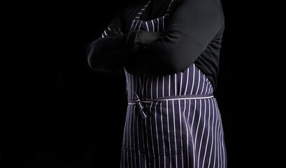  man chef in a striped blue apron and black clothes stands against a black background, his arms are crossed on his chest
