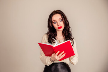Fototapeta premium a beautiful young girl with red lips holds a red notebook in her hands. student. businesswoman on isolated gray background