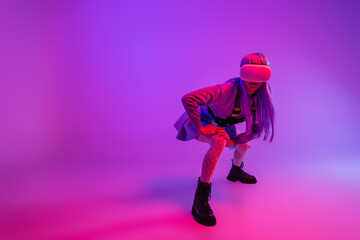 full length of young woman in virtual reality headset sitting on purple and pink