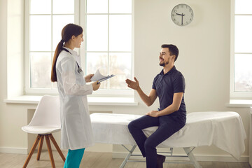 Fototapeta premium Happy patient talking to friendly doctor during health checkup at the hospital. Young man sitting on examination bed in office of modern clinic and asking physician questions about his treatment