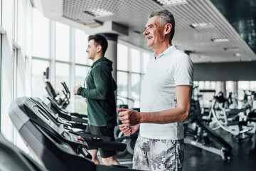 Fototapeta na wymiar Two men on a treadmill in the gym, in focus a gray-haired, smiling senior man. Sports, training, health. Sports Hall