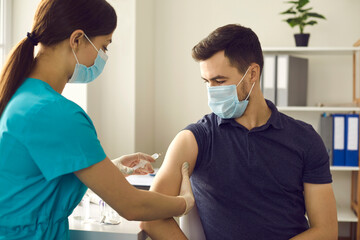 Professional nurse in medical face mask giving flu shot to male patient during seasonal vaccination campaign. Doctor injecting young man with influenza, AIDS or Covid-19 vaccine at clinic office - obrazy, fototapety, plakaty