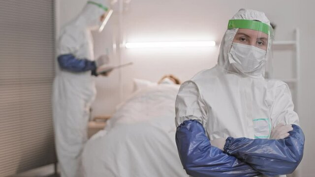 Waist-up POV of brown-eyed doctor wearing white hazmat suit, face shield and mask standing in hospital room, crossing arms on chest and looking on camera, colleague working on background