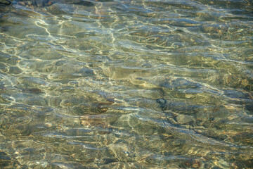 Meditative ripple of mountain lake. Beautiful relaxing background of stony bottom in transparent water of glacial lake in sunlight. Sunny backdrop with many stones in clear water of glacier lake.