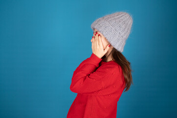 Side view of brunette girl in winter hat covering her face with her hands. Young woman in red...