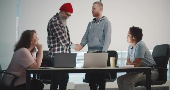 Hipster senior boss shaking hand of young male employee at team meeting in office