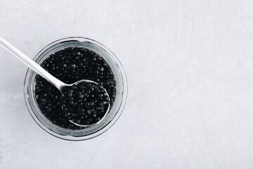 Fototapeta na wymiar Black caviar in glass jar with spoon on white stone background, luxurious delicacy appetizer.