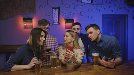 Friends in pub at bar counter with beer look at their phones. woman shows something cool on her phone to the friends. Social life