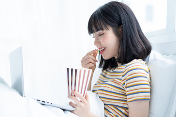 Young asian woman using laptop to watching tv shows, eating popcorn at home