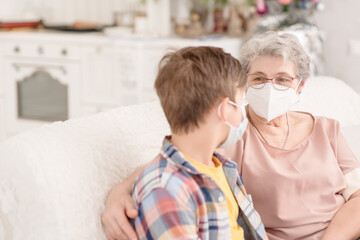 A grandson and an elderly woman with medical masks on their faces are sitting at home on the couch and chatting. Quarantine during coronavirus pandemic concept