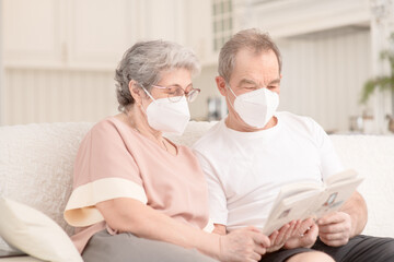 An elderly couple with medical masks are sitting on the couch at home and reading a book. Ways not to get bored during lockdown and develop while sitting at home. Quarantine during coronavirus pandemi