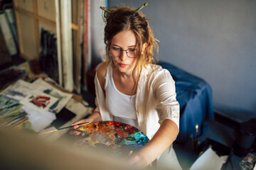 Artist female sitting next to the easel with canvas painting something in her art studio. A professional candid young woman painter in transparent eyeglasses draws in the workshop.