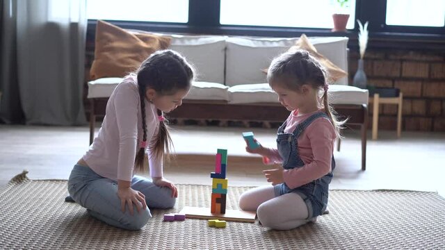 Happy sisters sitting on carpet, playing with colorful wooden blocks constructor