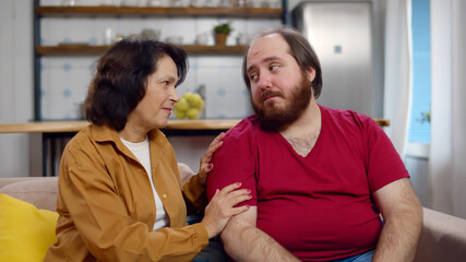 Senior woman comforting upset overweight adult son sitting together on couch