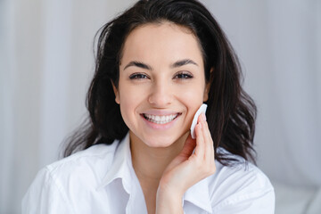 Smiling pretty young woman with clean skin using cotton pad in white comfortable bedroom