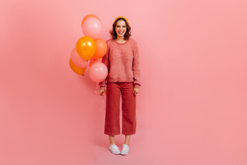 Full length view of smiling woman with air balloons. Charming curly girl posing with party decoration on pink background.