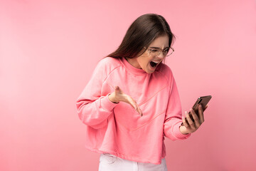 Photo of attractive lady in glasses holds telephone and screams, disappointed reads comments on blog. Wears casual pink t-shirt white pants isolated pink color background.