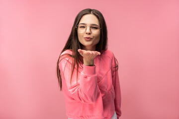 Photo of attractive young woman in glasses blows a kiss, wears casual pink t-shirt white pants...
