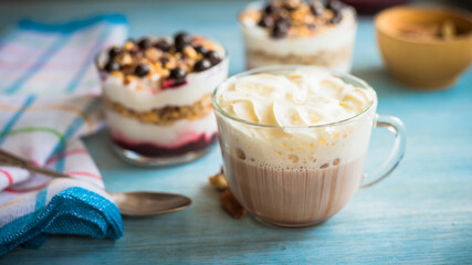Cup of cocoa with milk and a cap of whipped cream and creamy dessert in cups, spoon and napkin on a blue wooden background - breakfast