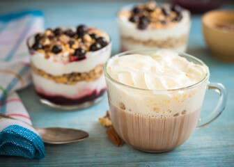 Cup of cocoa with milk and a cap of whipped cream and creamy dessert in cups, spoon and napkin on a blue wooden background - breakfast