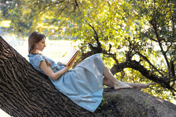 Young girl sitting on a tree by the river, reading a book. Copy space