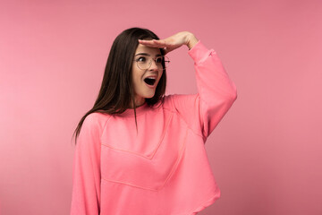 Fototapeta premium Photo of attractive young woman in glasses surprised, look into the distance. Wears casual pink t-shirt white pants isolated pink color background