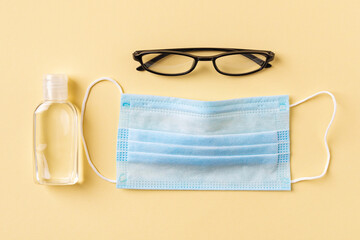 Disposable medical face mask, hand sanitizer in a small bottle and glasses on a yellow background....