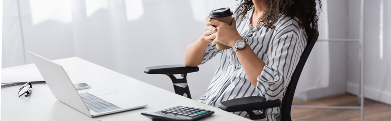 cropped view of african american freelancer holding paper cup near laptop on desk, banner