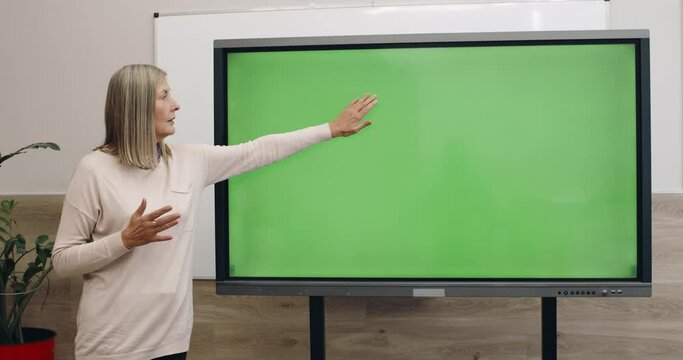 Smart woman talking while using and touching interactive screen with template green screen. Female professor explaining material and looking to camera while standing near digatal board.