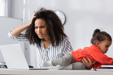stressed african american mother adjusting hair and looking at laptop near toddler kid crawling on desk