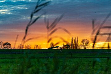 Fototapeta premium Behind the reeds along lake Zoetermeerse Plas, a cyclist cycles under a sky that takes on magical colors due to the setting sun and in the background the covered ski slope of Snowworld