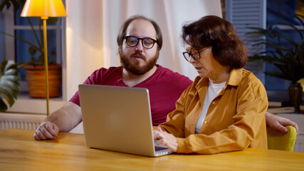 Young man helping mother using laptop sitting at table in living room