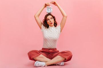 Curious girl with clock sitting on floor. Interested young woman posing on pink background. - obrazy, fototapety, plakaty