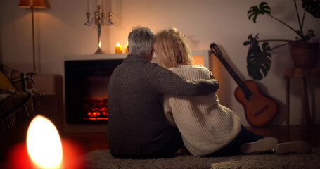Back view of aged husband and wife hugging relaxing on carpet near fireplace