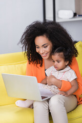smiling african american freelancer using laptop near toddler daughter in living room