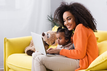 smiling african american mother using laptop near toddler daughter in living room