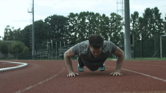Male athlete doing stratching before traning. He doing jumps and push ups on running track on stadium. Active lifestyle and determination. Strong, serious look and focus. Slow motion shot.