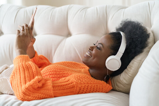 African millennial woman in orange sweater wearing wireless headphones, listening music, holding and using smart phone, resting and lying on couch in living room. Girl spend lazy weekend at home.