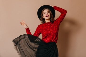 Enthusiastic girl touching hat with smile. Studio shot of excited caucasian woman dancing on brown...
