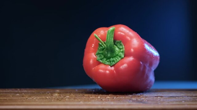 Red bell pepper with thick peduncle falls on brown wooden board reflecting bright electric light slow motion extreme closeup