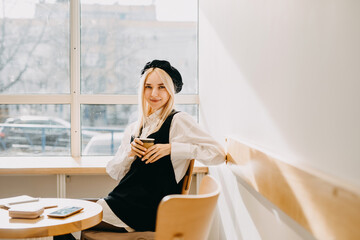 Young stylish woman sitting at a cafe table, holding a cup of coffee, smiling, looking at camera.