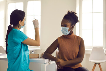 Professional nurse preparing flu vaccine injection for patient. Young woman in medical face mask getting COVID-19 shot and waiting for doctor to get syringe ready during vaccination in modern clinic - obrazy, fototapety, plakaty