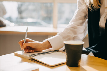 Closeup of a woman sitting at a table with a cup of coffee, writing down ideas in a notebook.