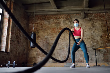 Young woman wearing face mask exercising with battle ropes at the gym. Strong female athlete doing crossfit workout with battle rope during epidemic COVID-19