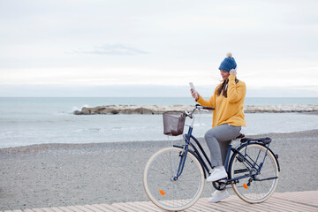 Fototapeta premium Beautiful active mature woman with bicycle on the road by the sea. Happy smiling middle aged woman with headphones listening to music, copy space