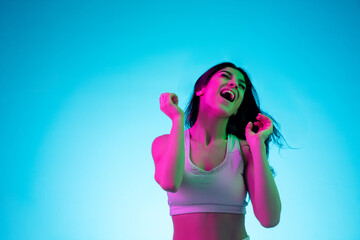 Laughting, dancing with hair blowed out. Brunette woman's portrait on blue studio background in mixed neon. Beautiful model in white top. Concept of human emotions, facial expression, sales, ad
