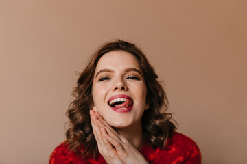 Lovable curly woman posing with tongue out. Studio shot of cheerful girl isolated on brown background.